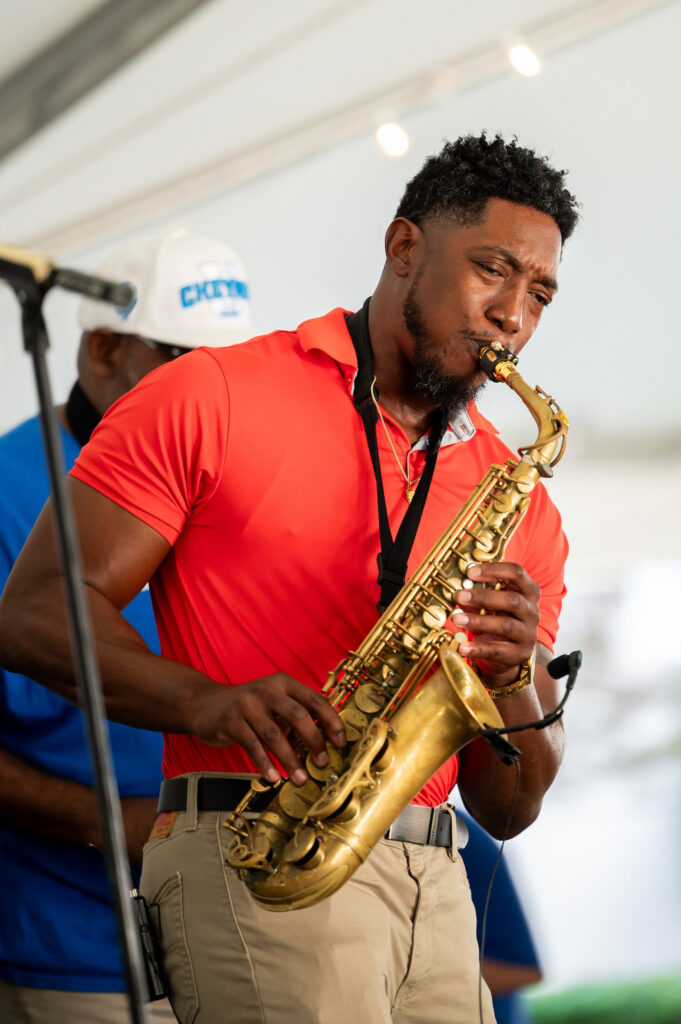 A person in a red shirt plays a golden saxophone on stage under a tent.