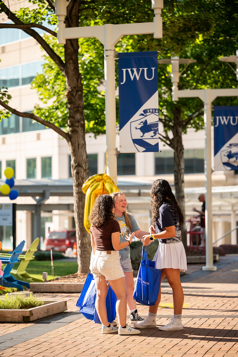 Three students laugh and chat on a sunny day at a university campus, surrounded by trees, banners, and colorful balloons.