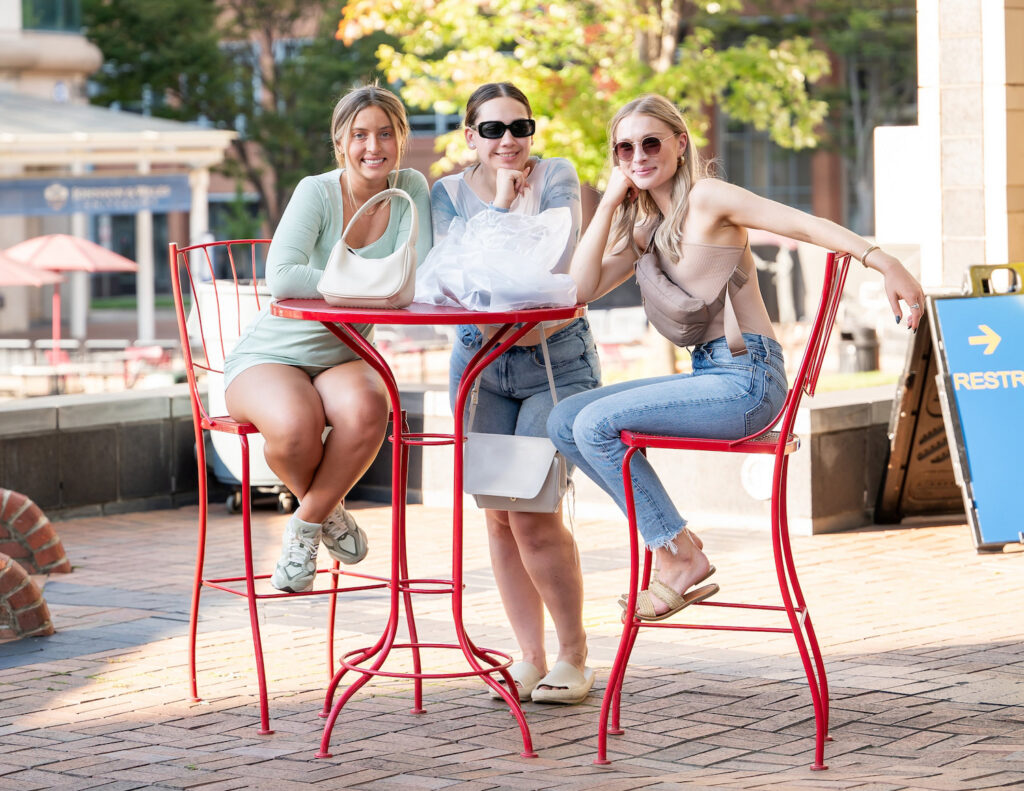 Three individuals sit at a red outdoor table on a brick-paved plaza, enjoying a sunny day with trees and buildings in the background.