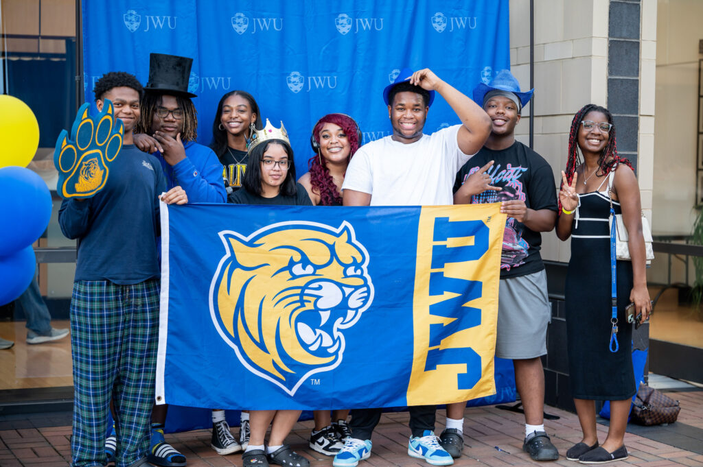 A group of students poses with a J.W.U. flag and mascot-themed accessories in front of a branded backdrop.
