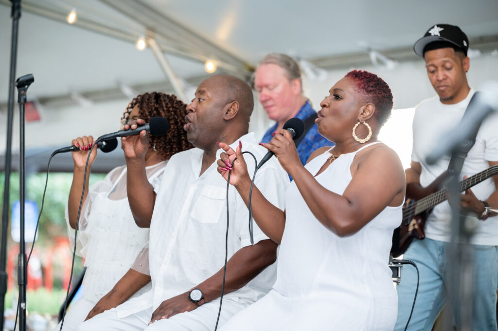 A group of performers dressed in white sings into microphones on a stage under a tent, with a guitarist visible in the background.