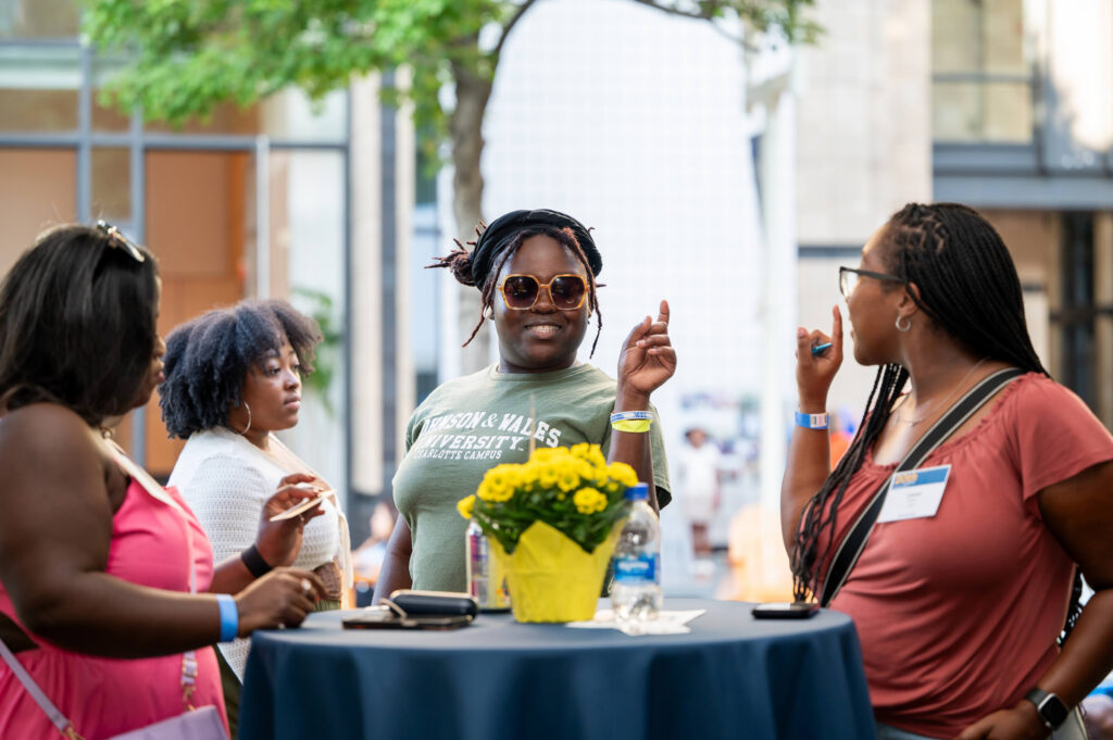 Four individuals engage in conversation around a table adorned with yellow flowers and beverages, set against an urban backdrop with trees and buildings.