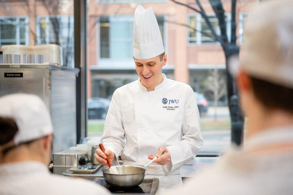 A chef in a white uniform and tall hat stirs a bowl while smiling, with students in chef's coats observing in a kitchen setting.