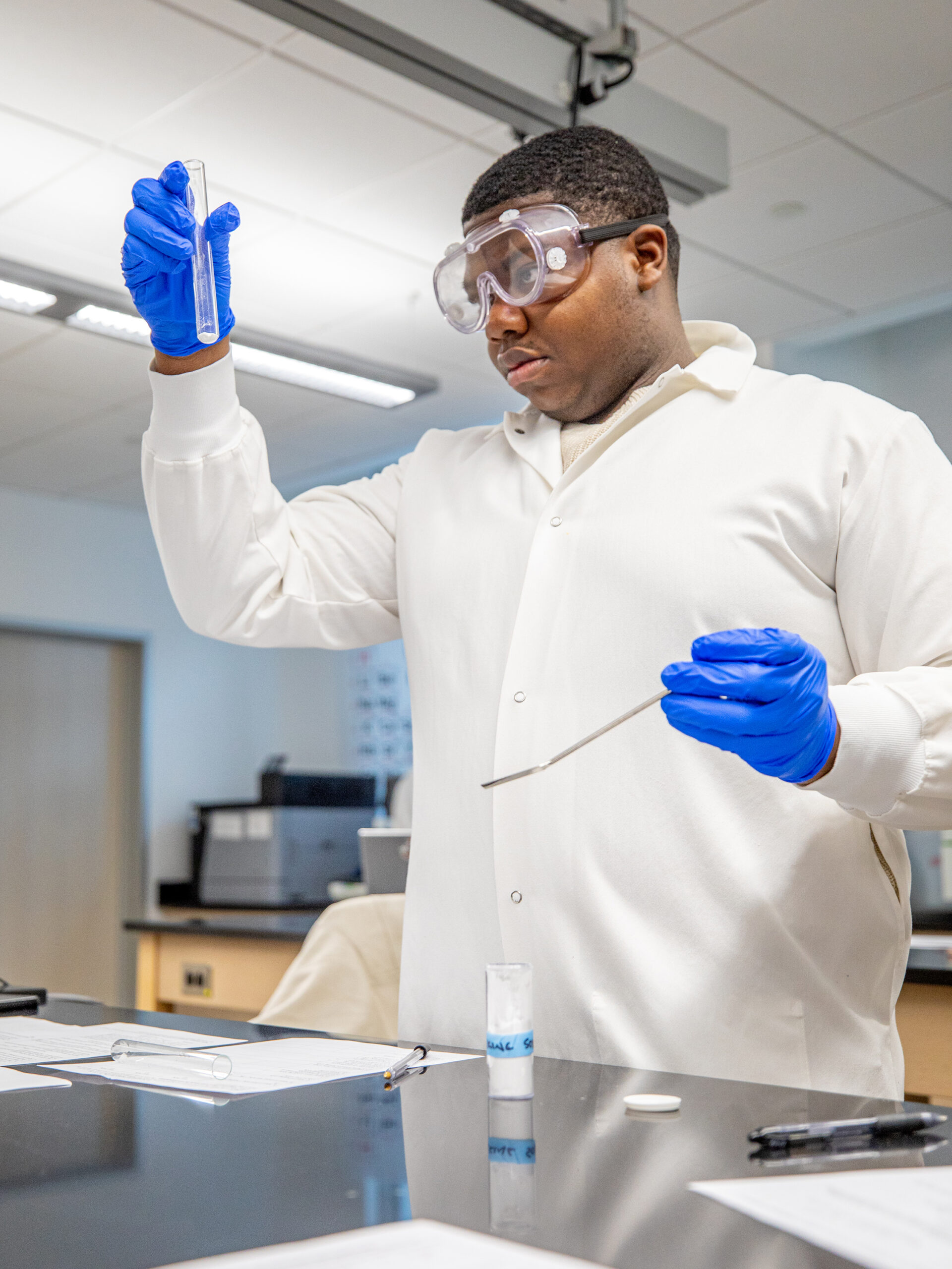 A scientist in a lab coat and goggles holds a test tube while using a tool to examine its contents.