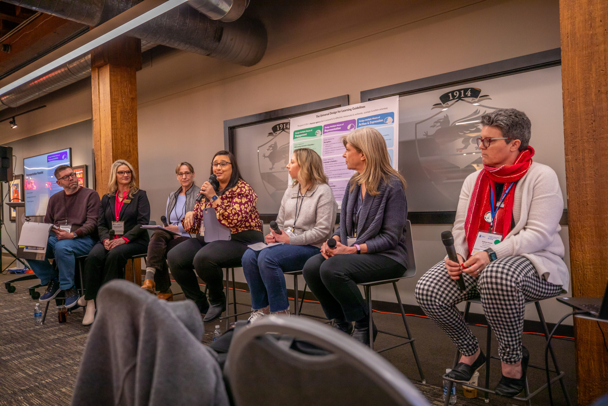 A panel of six individuals sits on stools in a conference room, with one person speaking into a microphone while others listen attentively.