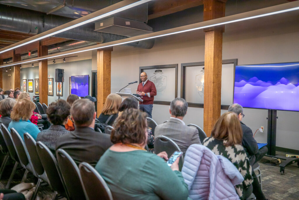 An individual speaks at a podium to an audience seated in chairs within a room featuring exposed wooden beams and large screens displaying abstract imagery.
