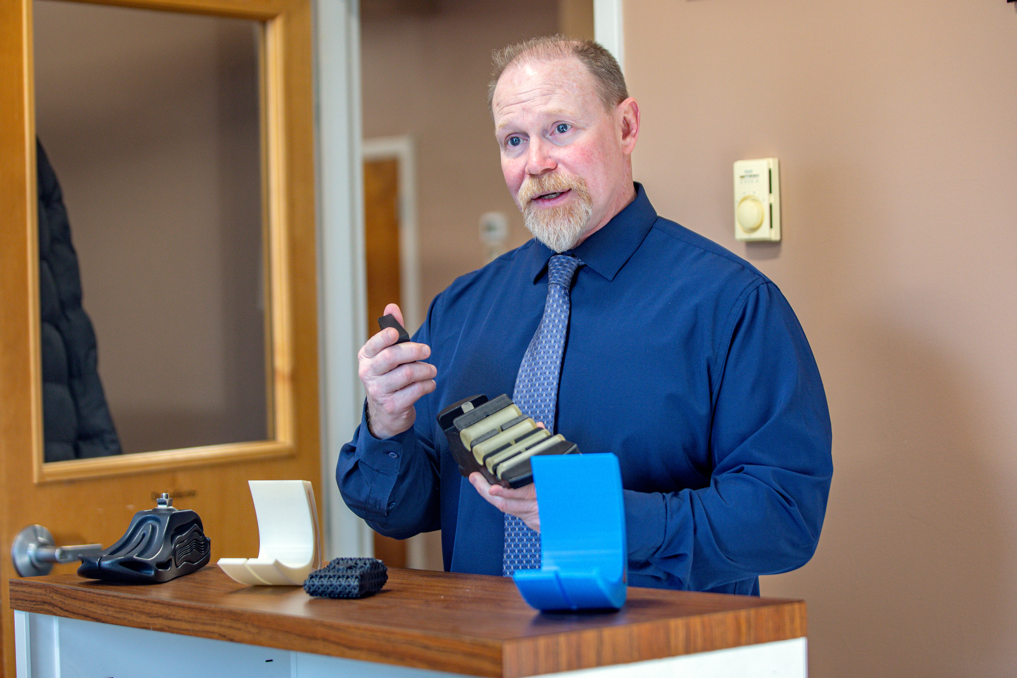 A person in a blue shirt and tie holds a small device while standing beside a counter displaying various 3D-printed objects.