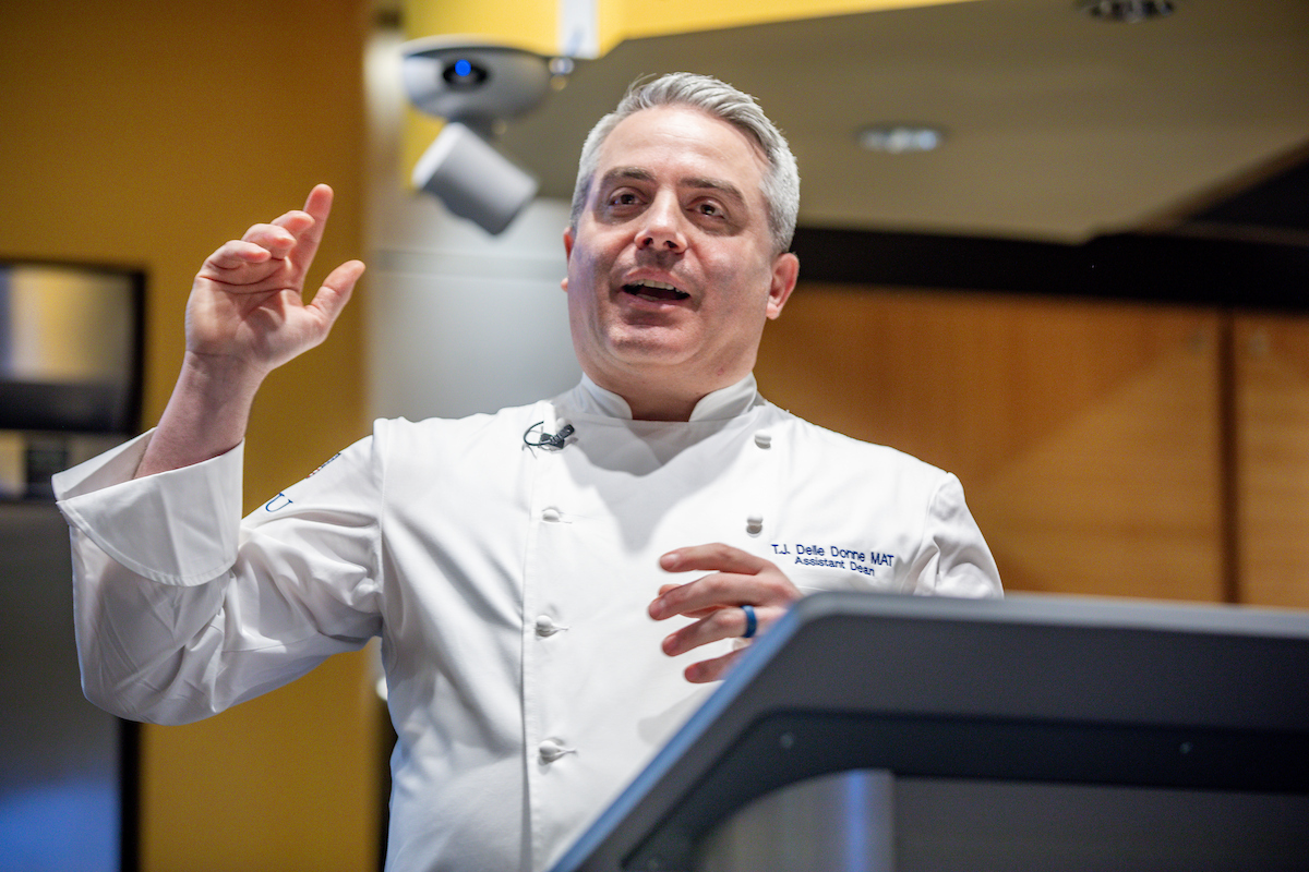 A chef in a white uniform gestures while speaking at a podium.