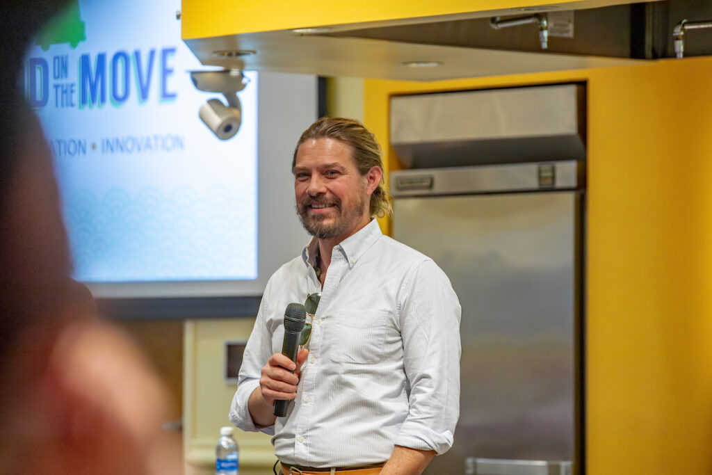 A person with long hair and a beard speaks into a microphone at an event, standing in front of a screen displaying “ON THE MOVE” and “INNOVATION.”
