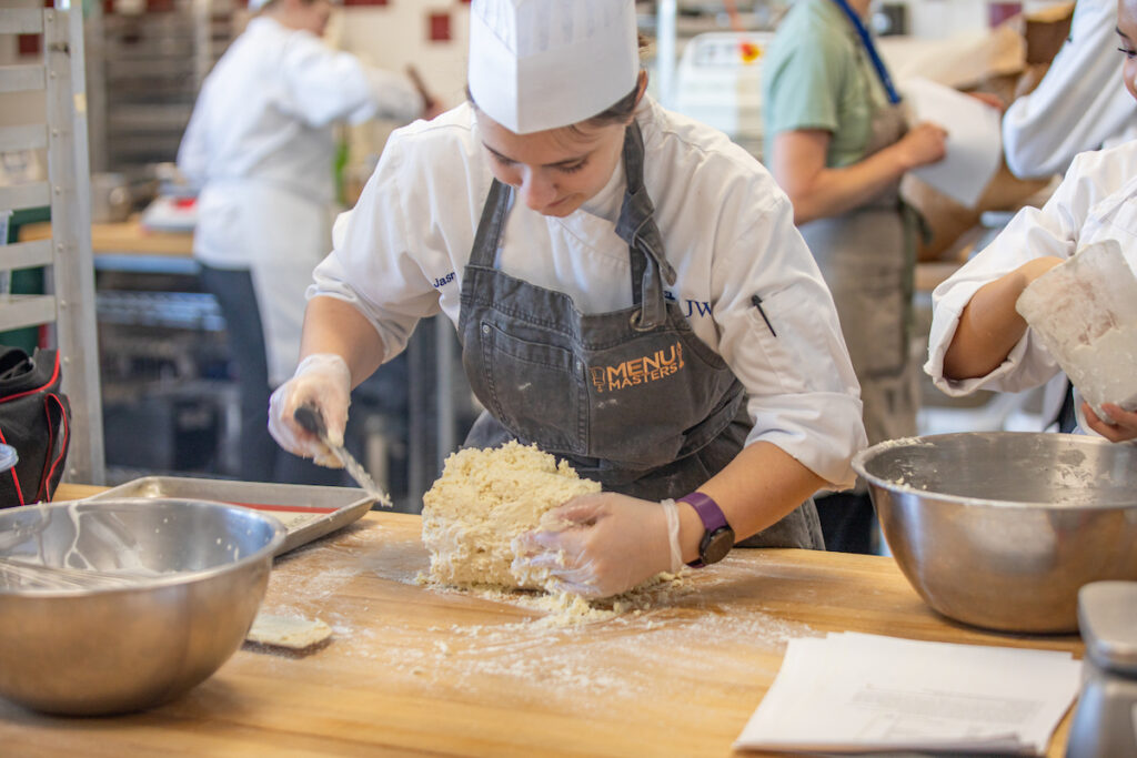 A chef in a kitchen kneads dough on a floured wooden surface while wearing a chef’s coat and apron.