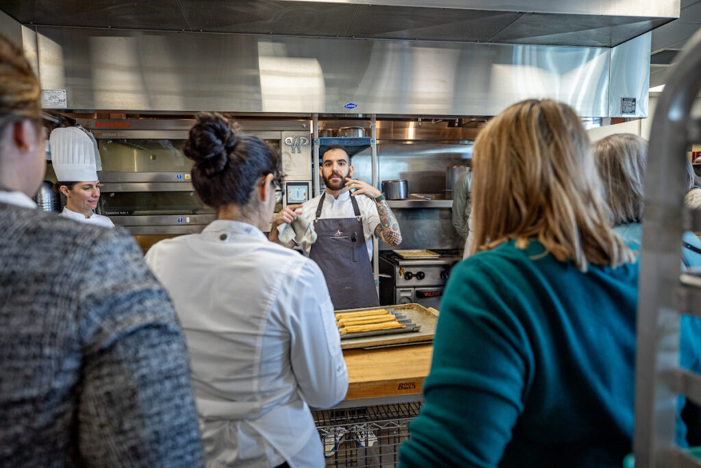 A chef demonstrates a cooking technique to a group of attendees in a professional kitchen setting.