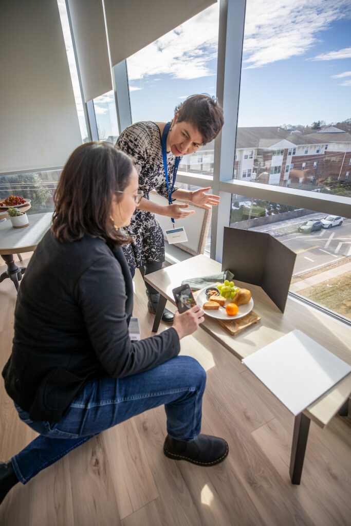 Two individuals collaborate indoors near a large window, one capturing a photo of a plated snack while the other gestures toward it.