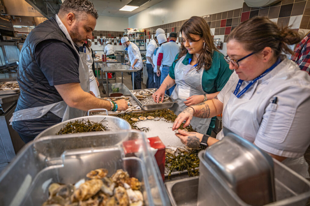 Several individuals in a kitchen prepare oysters and seaweed on a work surface, with others working in the background.