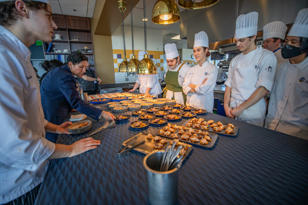 A group of chefs in a professional kitchen attentively observe and assist in plating an array of small dishes on a long counter.