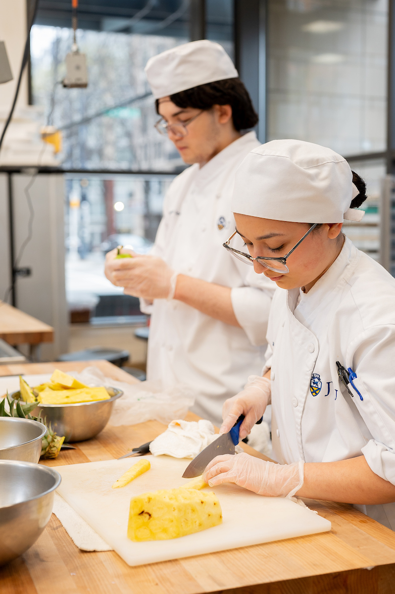 Two individuals in chef uniforms and gloves are preparing pineapple in a kitchen.