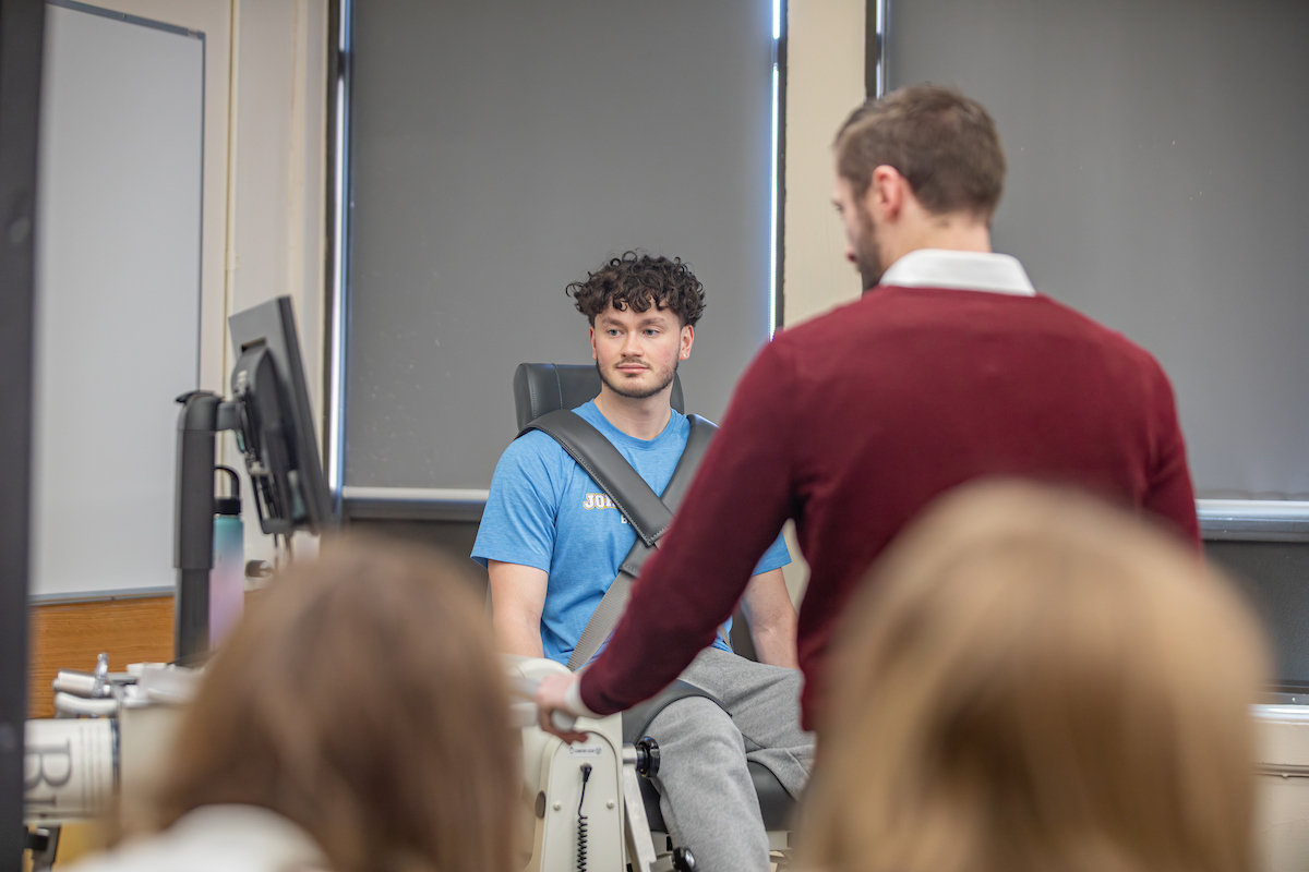 A student sits in a chair with safety harnesses on, looking at a monitor as the professor gives a lecture in a classroom.