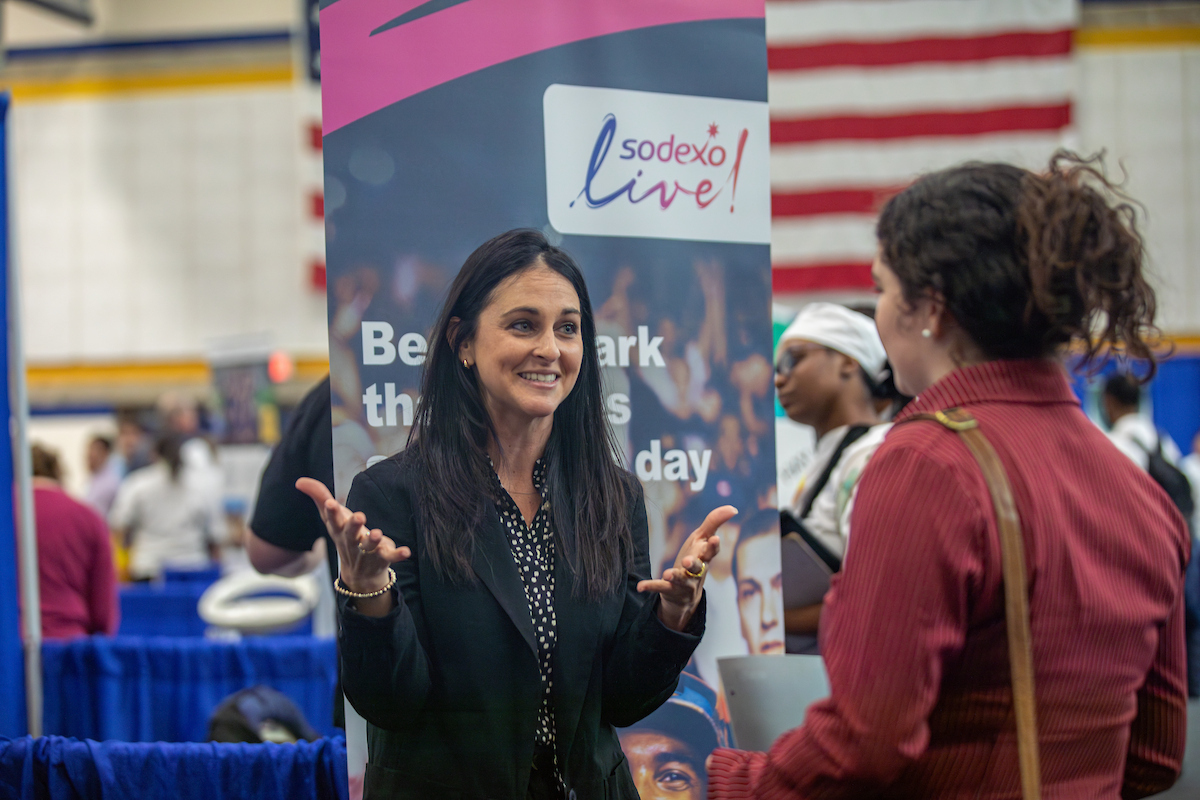 A person in a black blazer gestures while speaking to another individual at a Sodexo Live! event booth, with an American flag visible in the background.