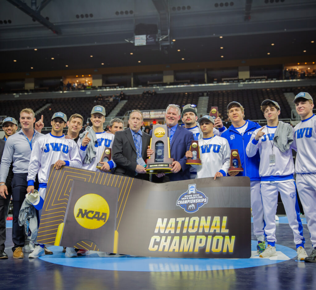 A wrestling team and staff pose on a mat with a large “NCAA National Champion” banner and trophies, celebrating their victory at a championship event.