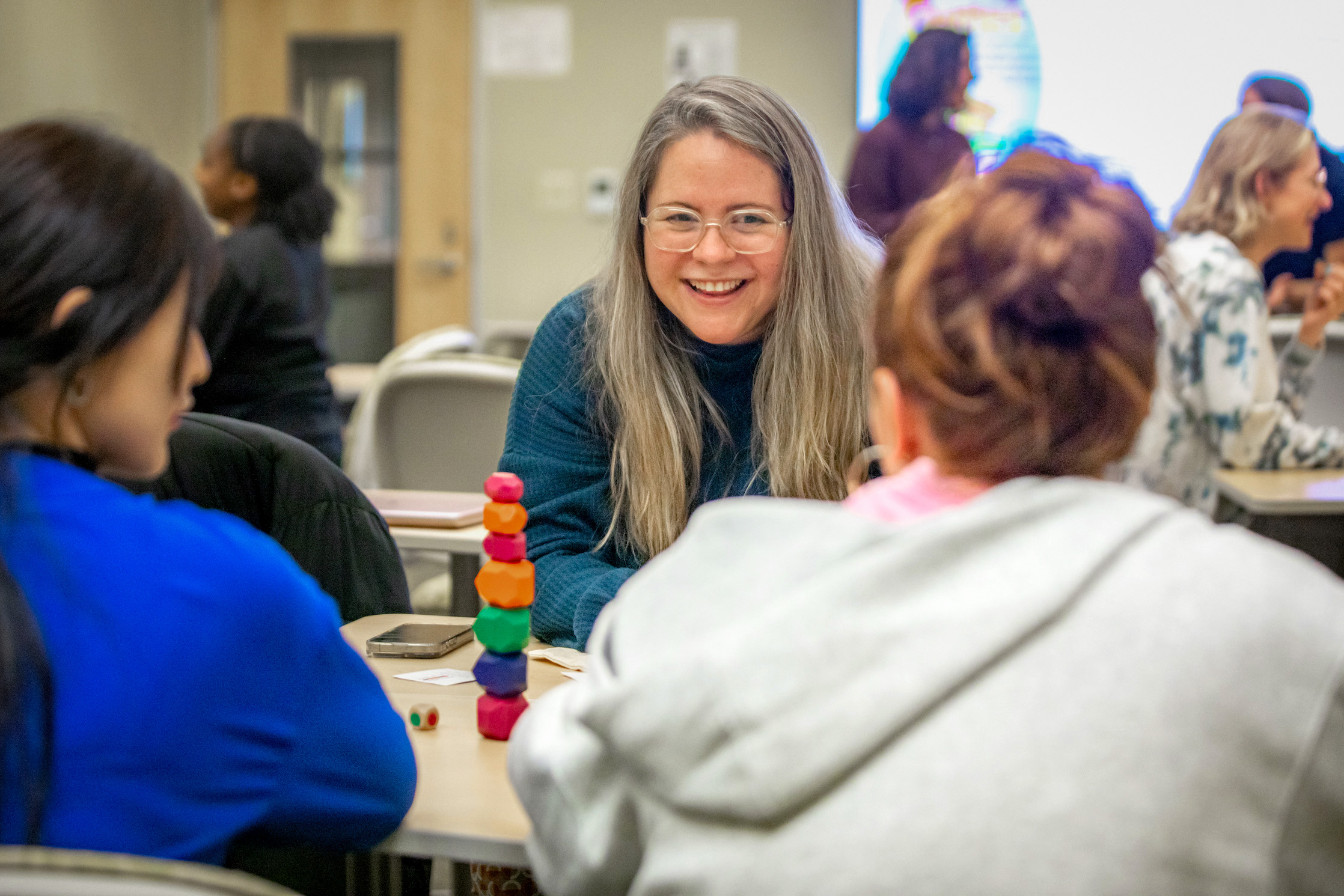 A smiling person with long hair and glasses engages in a tabletop game with others in a classroom setting.