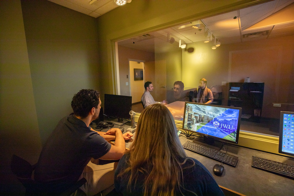 Several individuals are gathered around a desk in a room, observing a screen displaying a logo for JWU, while others are visible in an adjacent space.
