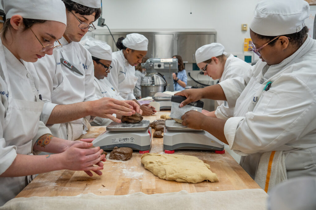 A group of culinary students in white uniforms and hats work together in a kitchen, carefully portioning dough on digital scales.