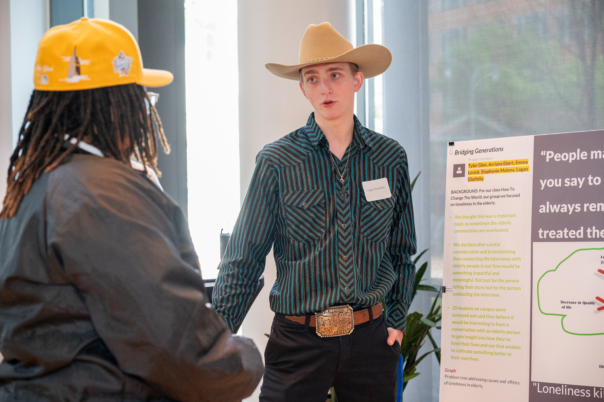 A person in a cowboy hat and striped shirt stands facing another person wearing a yellow cap, near a presentation board discussing loneliness among the elderly.