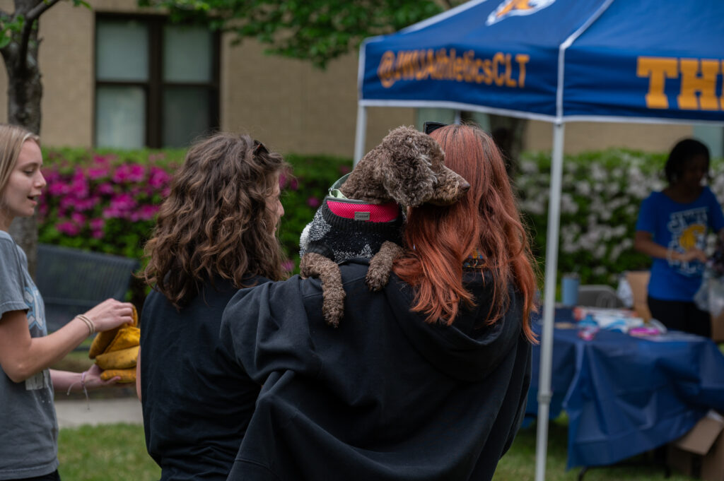 A person with long red hair carries a small brown dog on their shoulder at an outdoor event under a blue tent.