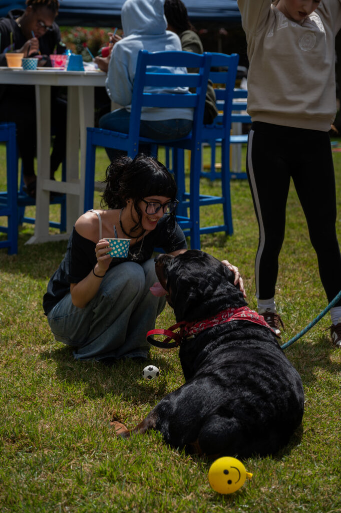 A person crouches on grass beside a large black dog, holding a cup and smiling, with toys nearby and others seated at tables in the background.