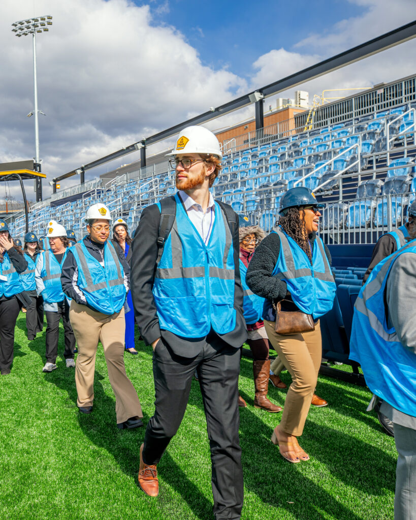 Students wearing hard hats and safety vests walk together through a stadium construction site.
