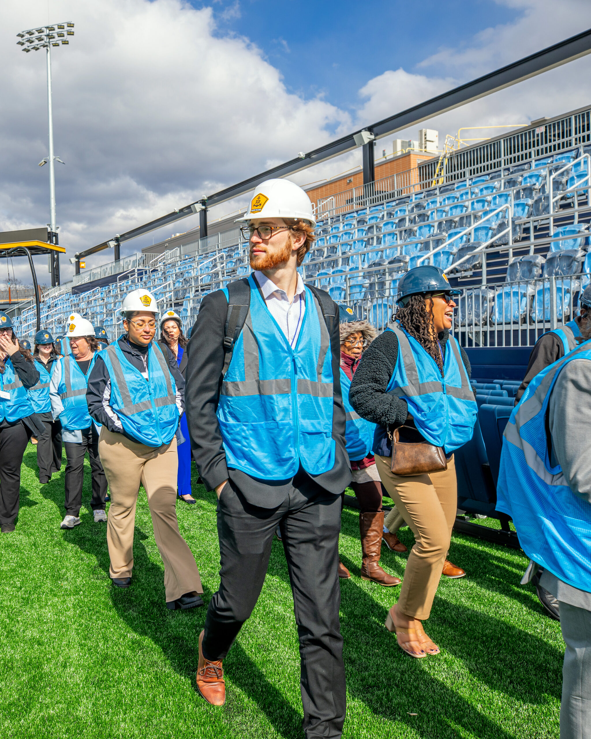 Students wearing hard hats and safety vests walk together through a stadium construction site.