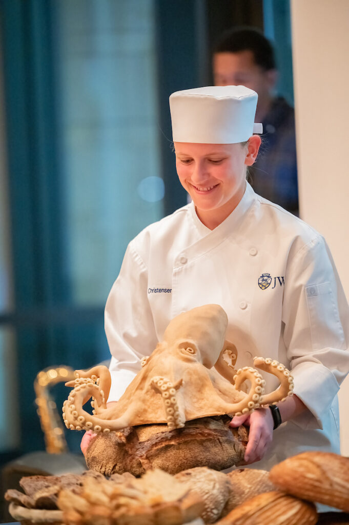 A chef in a white uniform presents a large, intricately crafted octopus sculpture made from bread, smiling as they hold it.