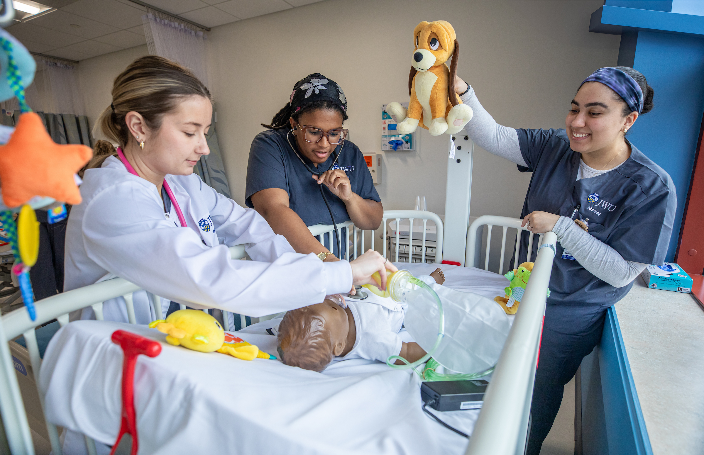 Three healthcare trainees work together with a baby mannequin in a pediatric simulation setting, one using a stethoscope, another adjusting an oxygen mask, and the third holding a stuffed animal.