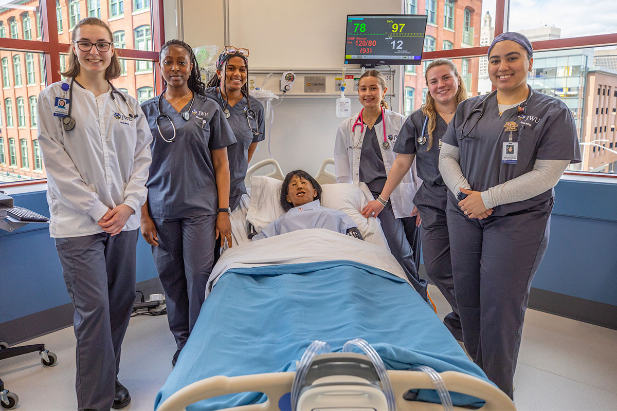 A group of nursing students in scrubs stand around a hospital bed with a mock patient, smiling for the camera in a brightly lit room with large windows.