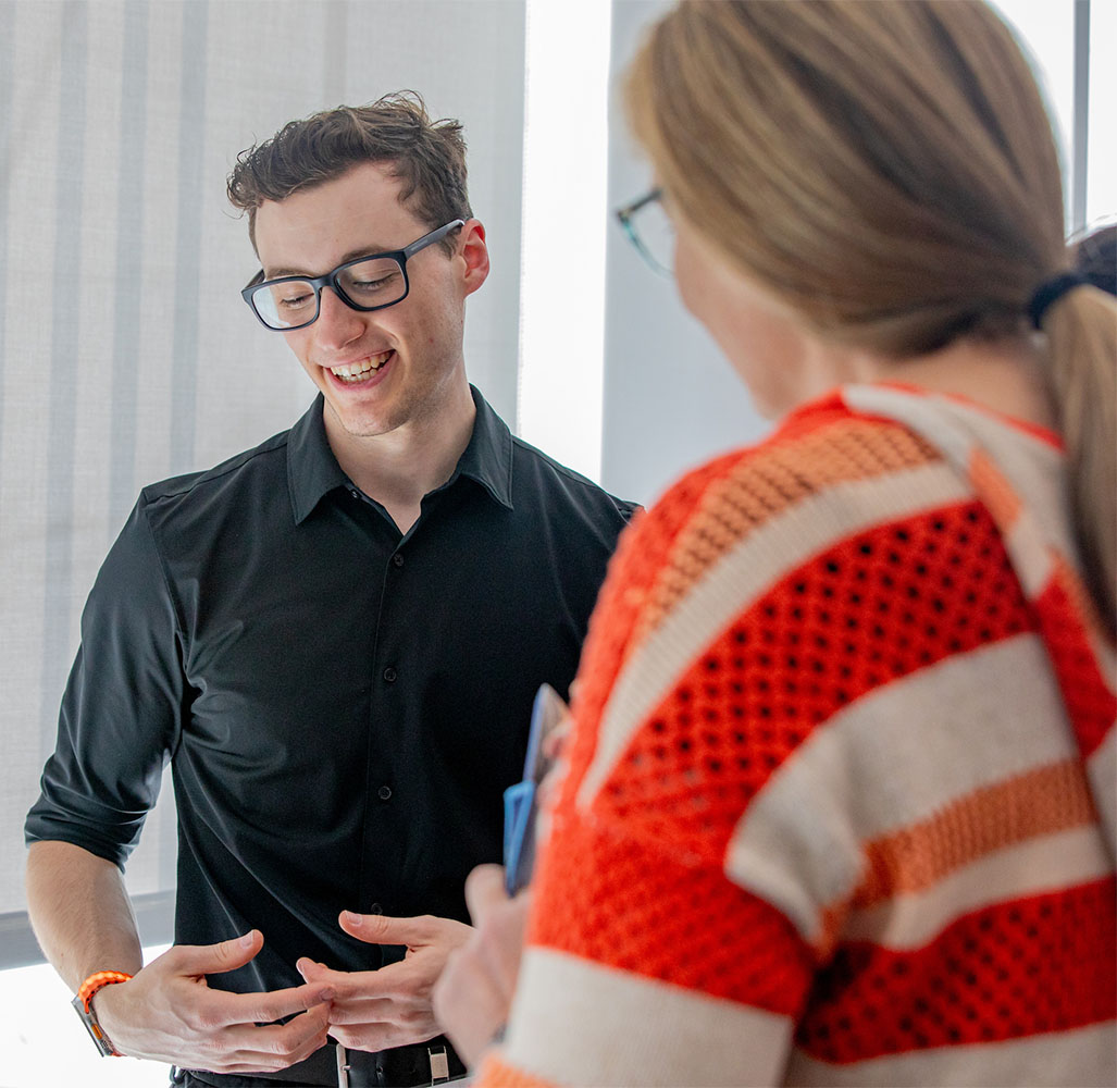 A student in a black shirt and glasses smiles while engaging in conversation with another person.
