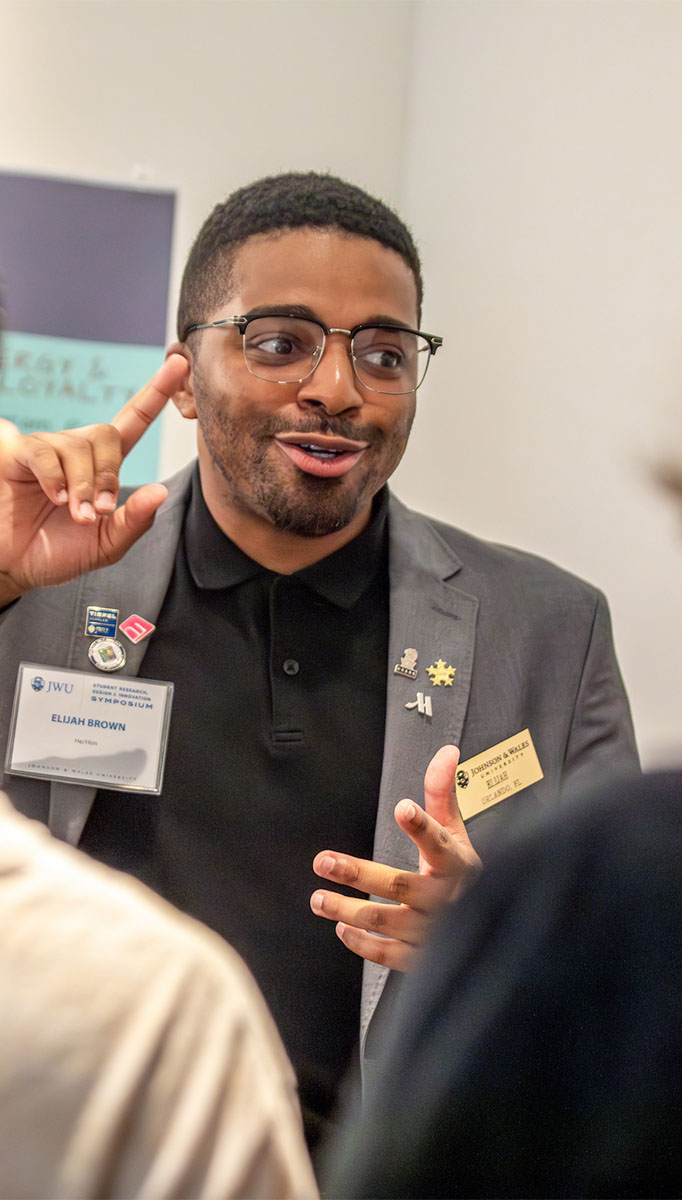 A student in a suit and glasses gestures while speaking to a group of attendees.