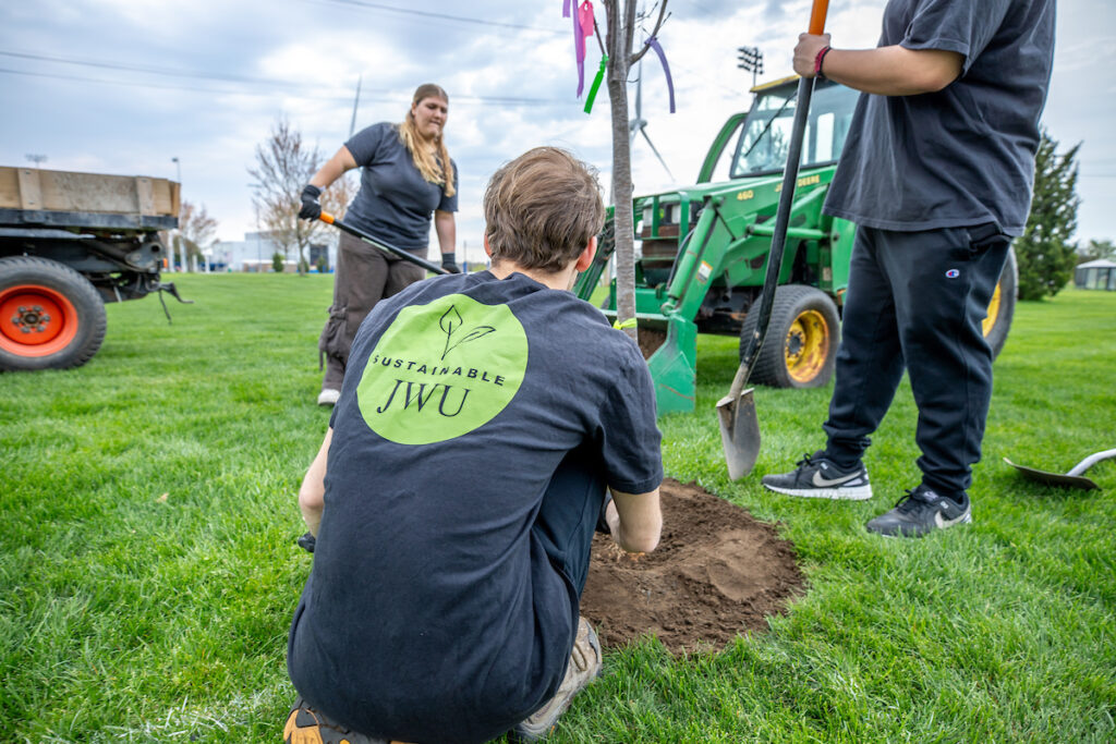 Individuals participate in a tree-planting event, with one person kneeling in the dirt while others stand nearby with shovels and a tractor in the background.