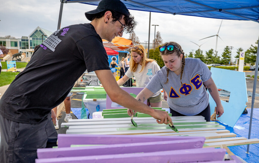 Several individuals work together under a blue canopy, painting wooden panels with green and purple paint.