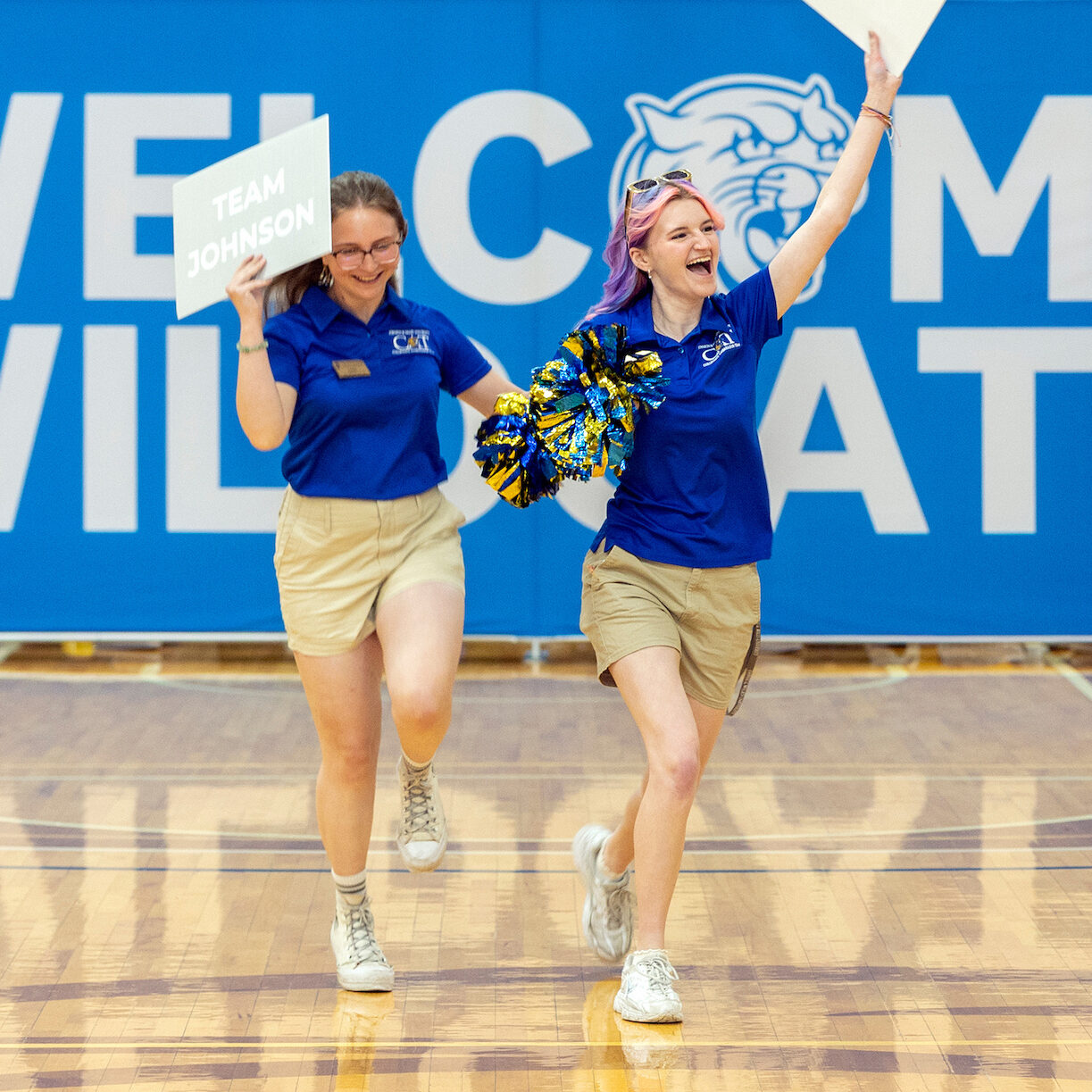 Two people cheer and hold signs in front of a large Welcome Wildcats banner.