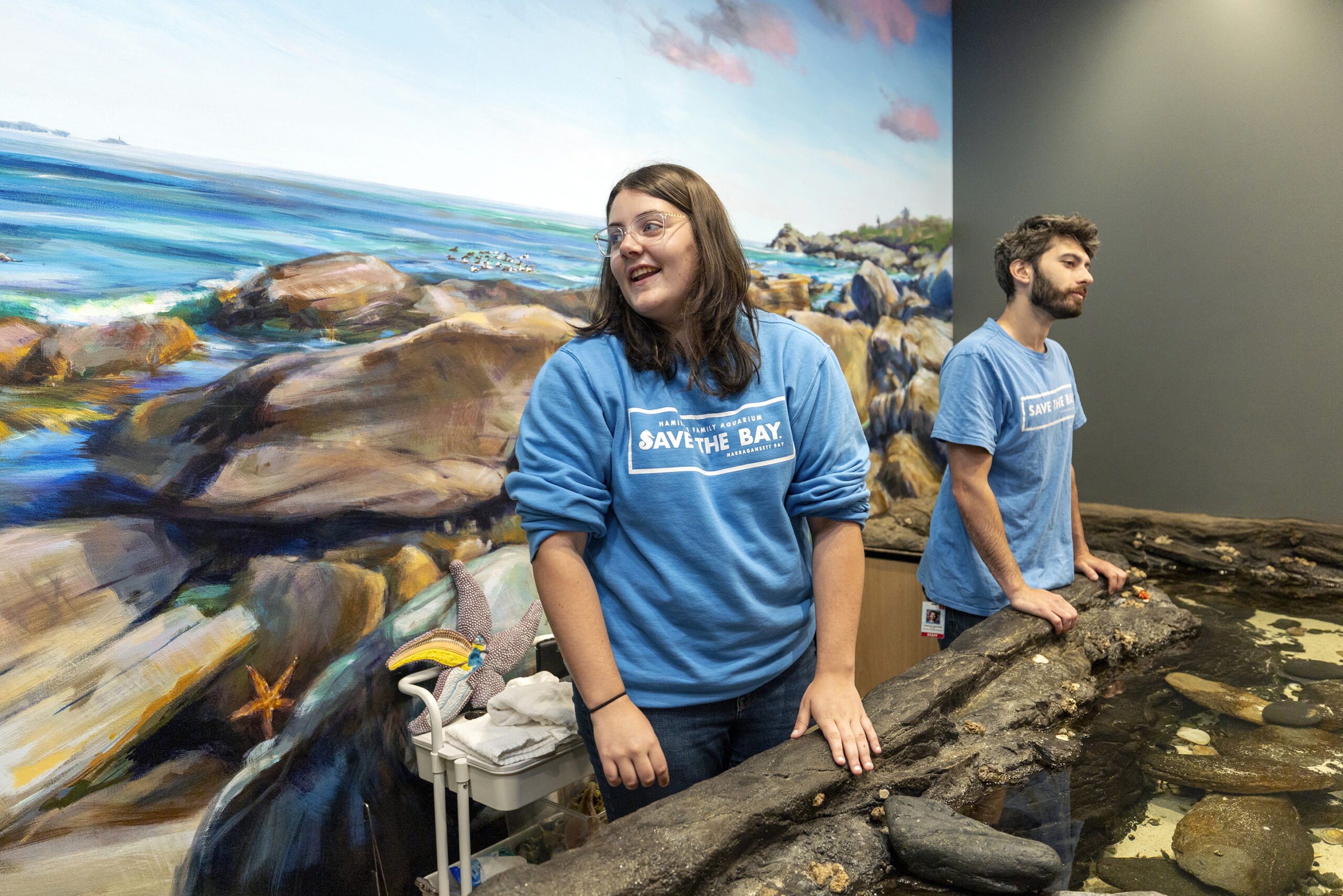Two individuals in blue "Save the Bay" shirts stand beside a rocky tide pool display, with a large ocean mural behind them.