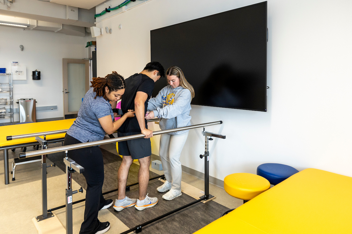 Three individuals assist a person with balance training using parallel bars in a clinical setting.