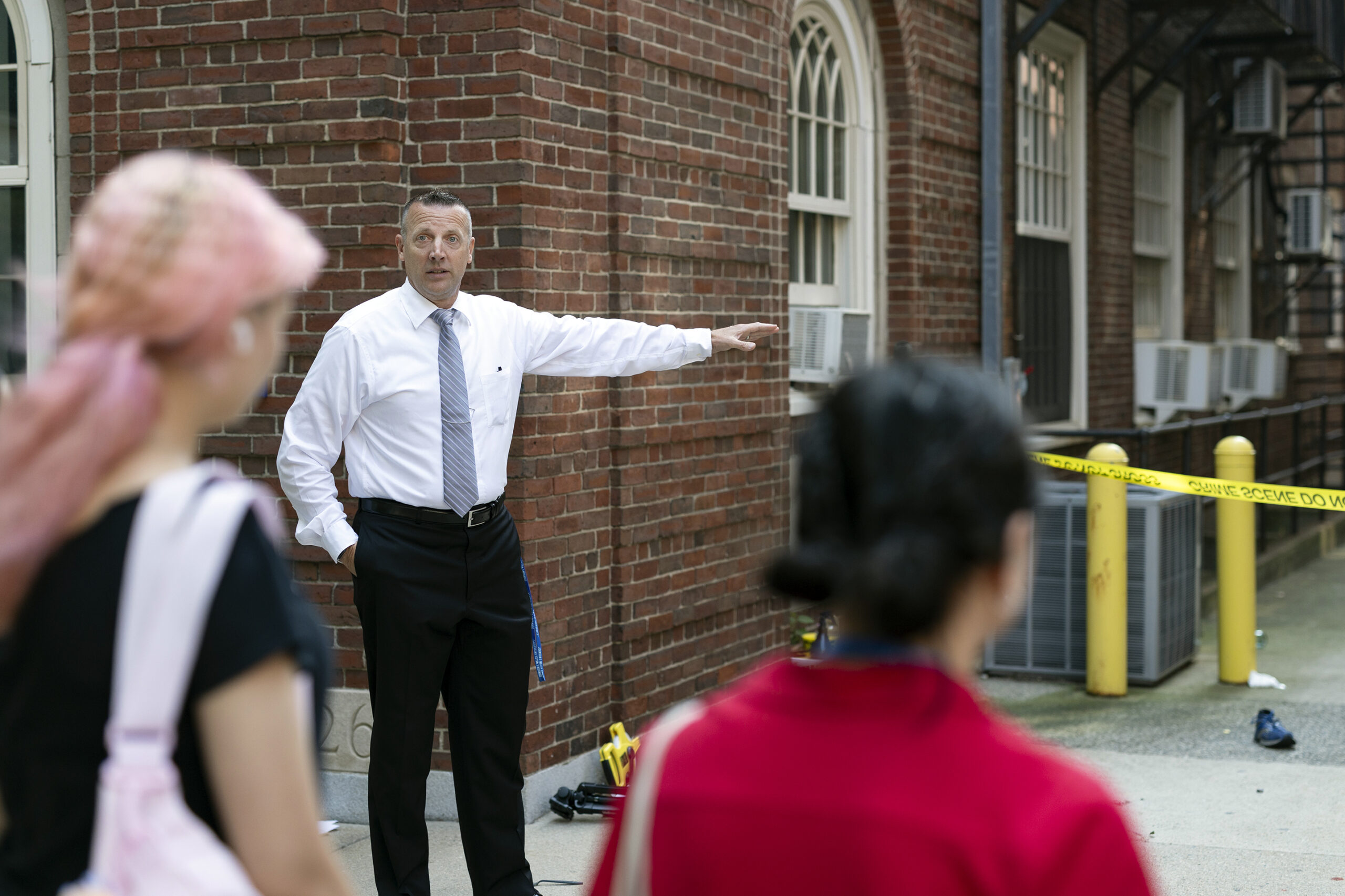 A person in a white shirt and tie gestures toward a brick building while others stand nearby, with yellow caution tape marking an area in the background.