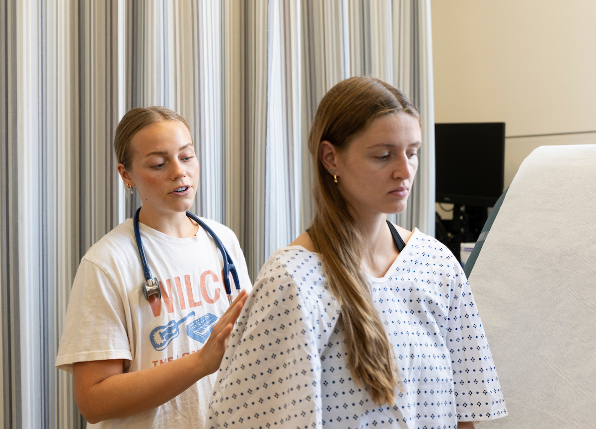 A healthcare worker with a stethoscope gently places a hand on another person’s shoulder while they both look downward.