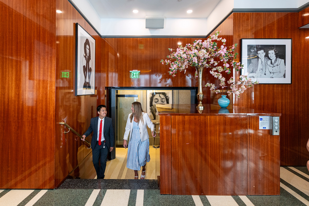 Two individuals walk through a warmly lit lobby with polished wood paneling, a reception desk adorned with floral arrangements, and framed artwork on the walls.