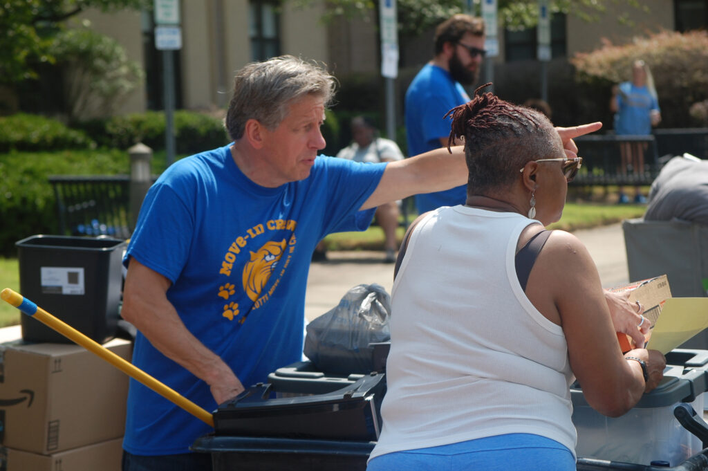 A person in a blue shirt points while another handles items near bins outdoors.