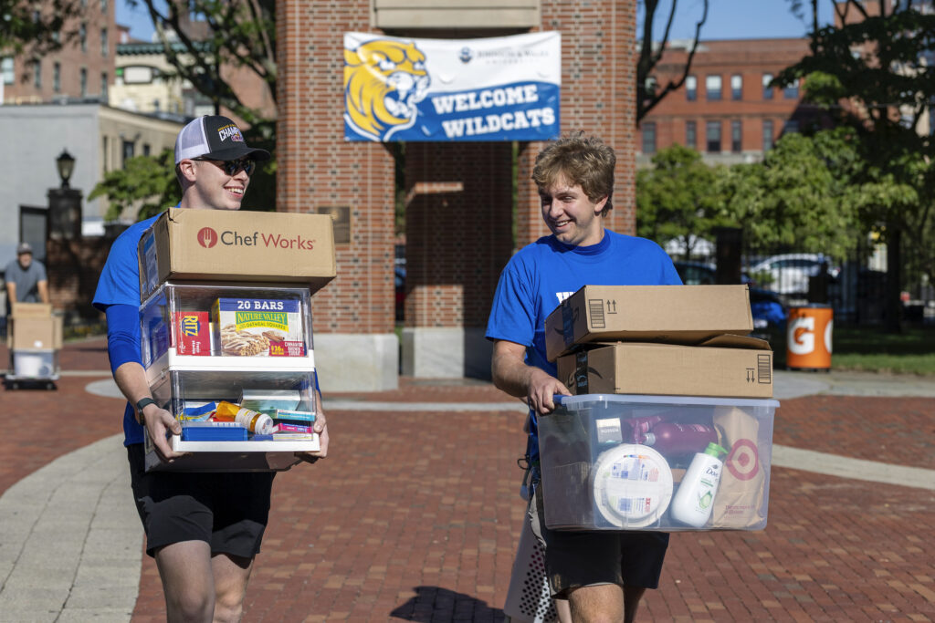 Two individuals carry boxes and containers of groceries and supplies across a brick-paved area near a university entrance with a "Welcome Wildcats" banner.