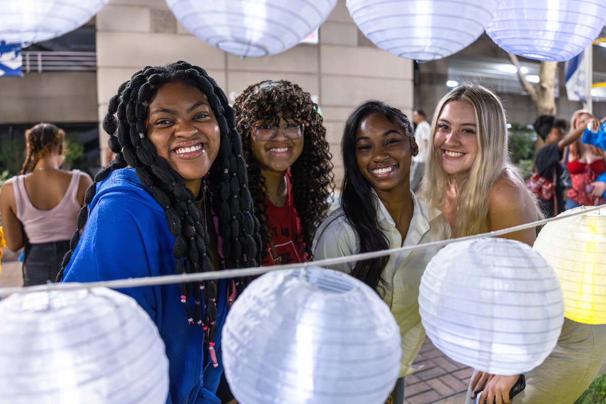Four smiling individuals pose together under illuminated paper lanterns at an outdoor evening event.