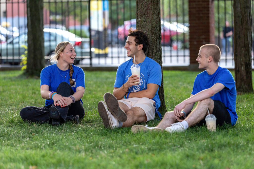 Three students sitting and talking on the grass in a shaded campus area.