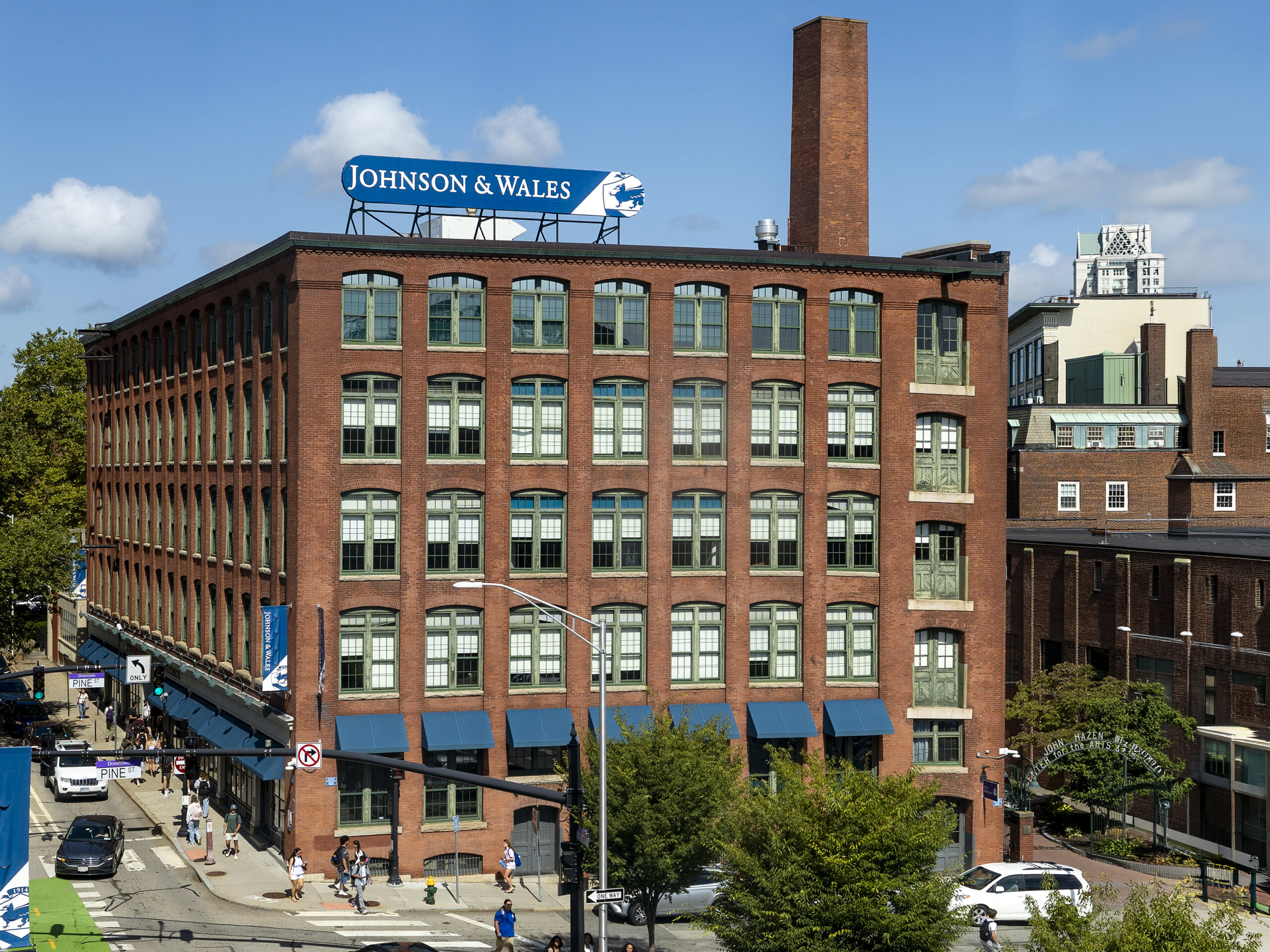 A red brick building with a "Johnson & Wales" sign overlooks a bustling street with pedestrians, vehicles, and trees under a clear blue sky.