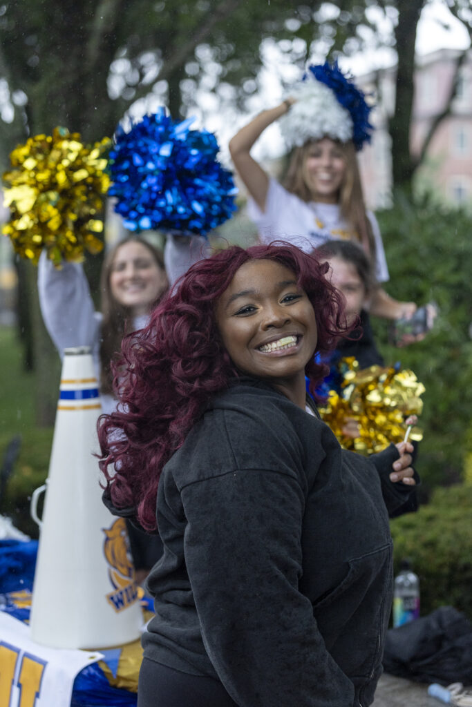 A person with vibrant red curls smiles brightly while holding a lollipop, surrounded by cheerleaders waving colorful pom-poms outdoors.