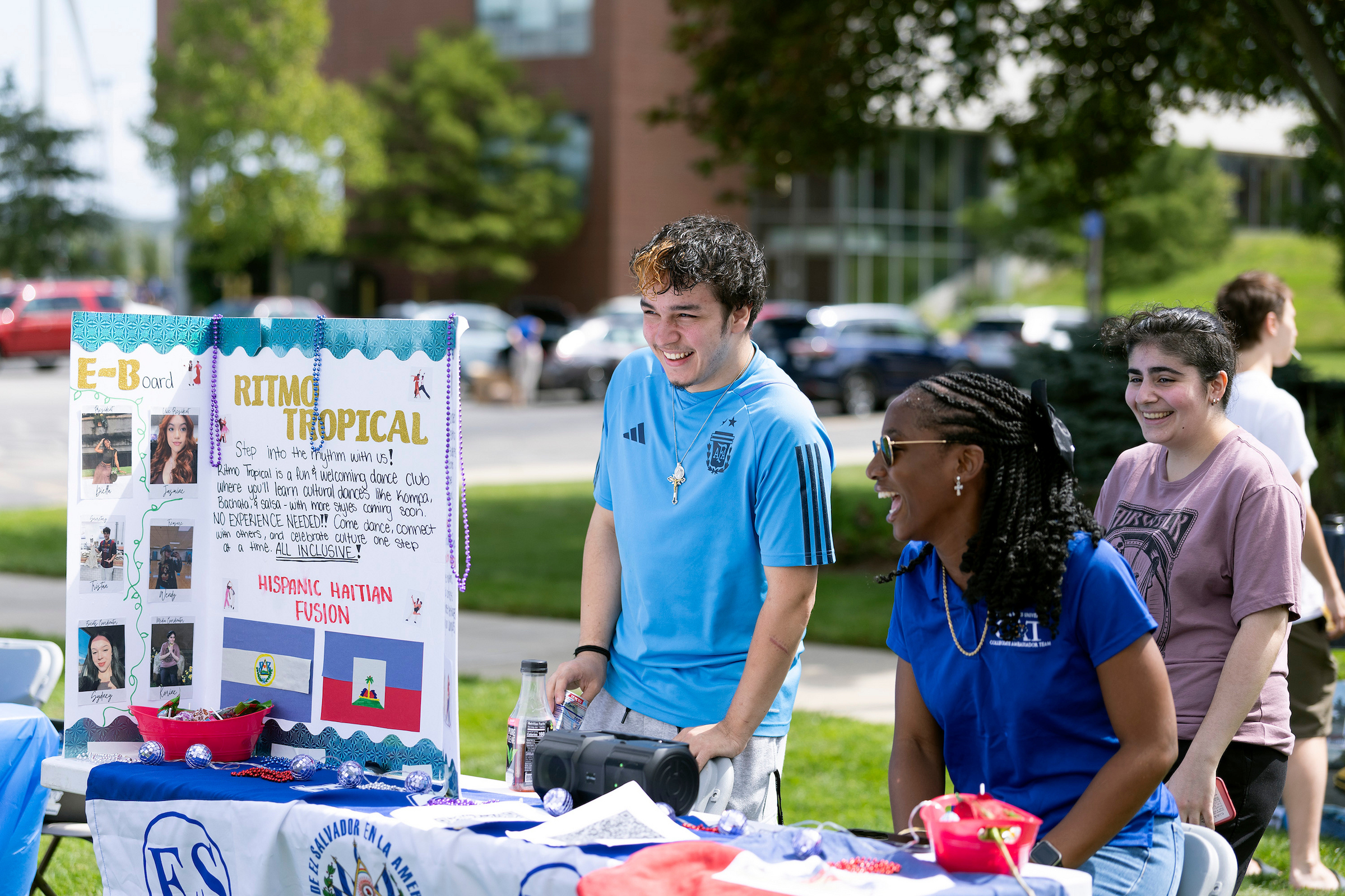 Three individuals engage with a colorful outdoor booth promoting a tropical dance event, surrounded by festive decorations and informational posters.